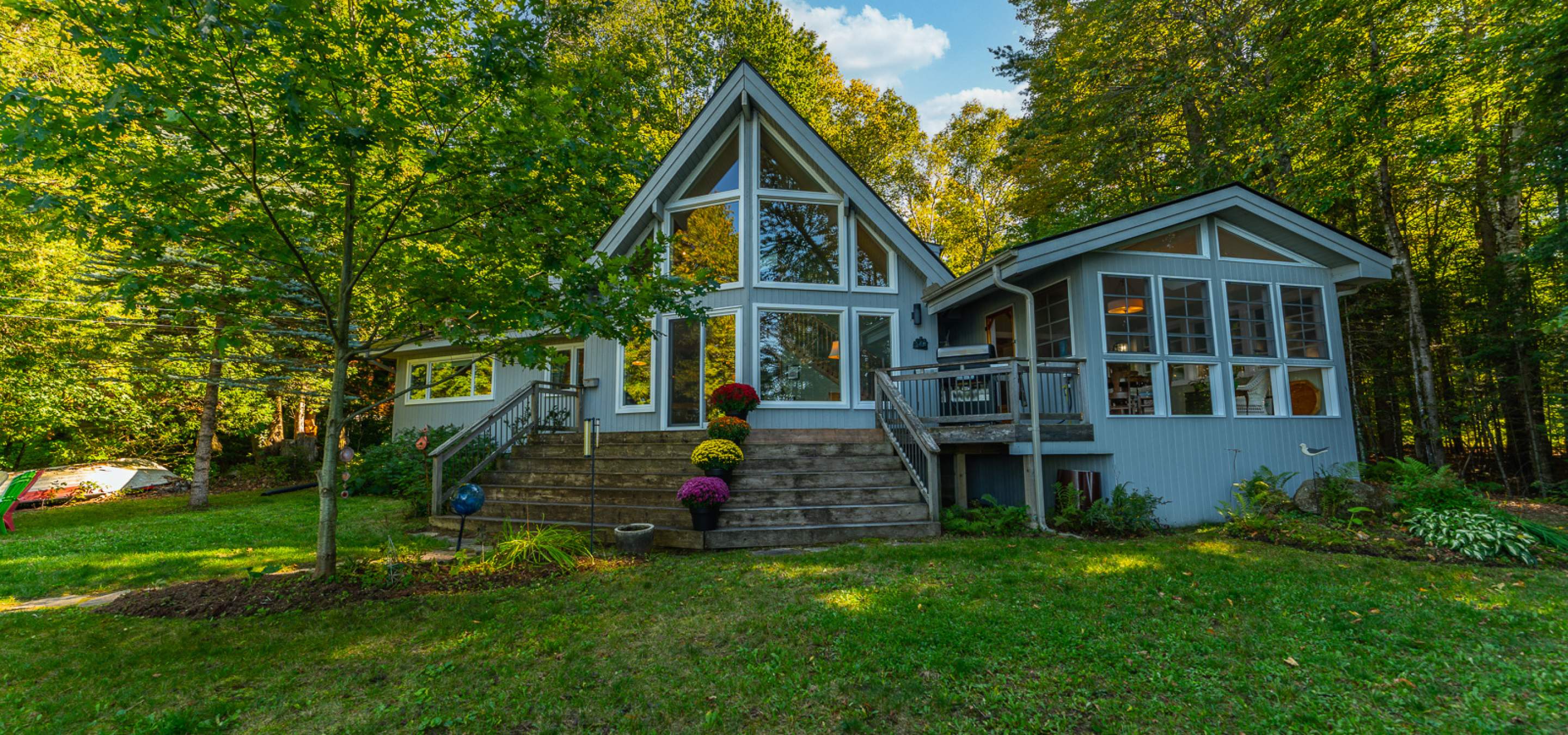 A charming lakeside home with large A-frame windows, a wooden deck, and vibrant flowers, nestled in a serene, tree-filled setting.